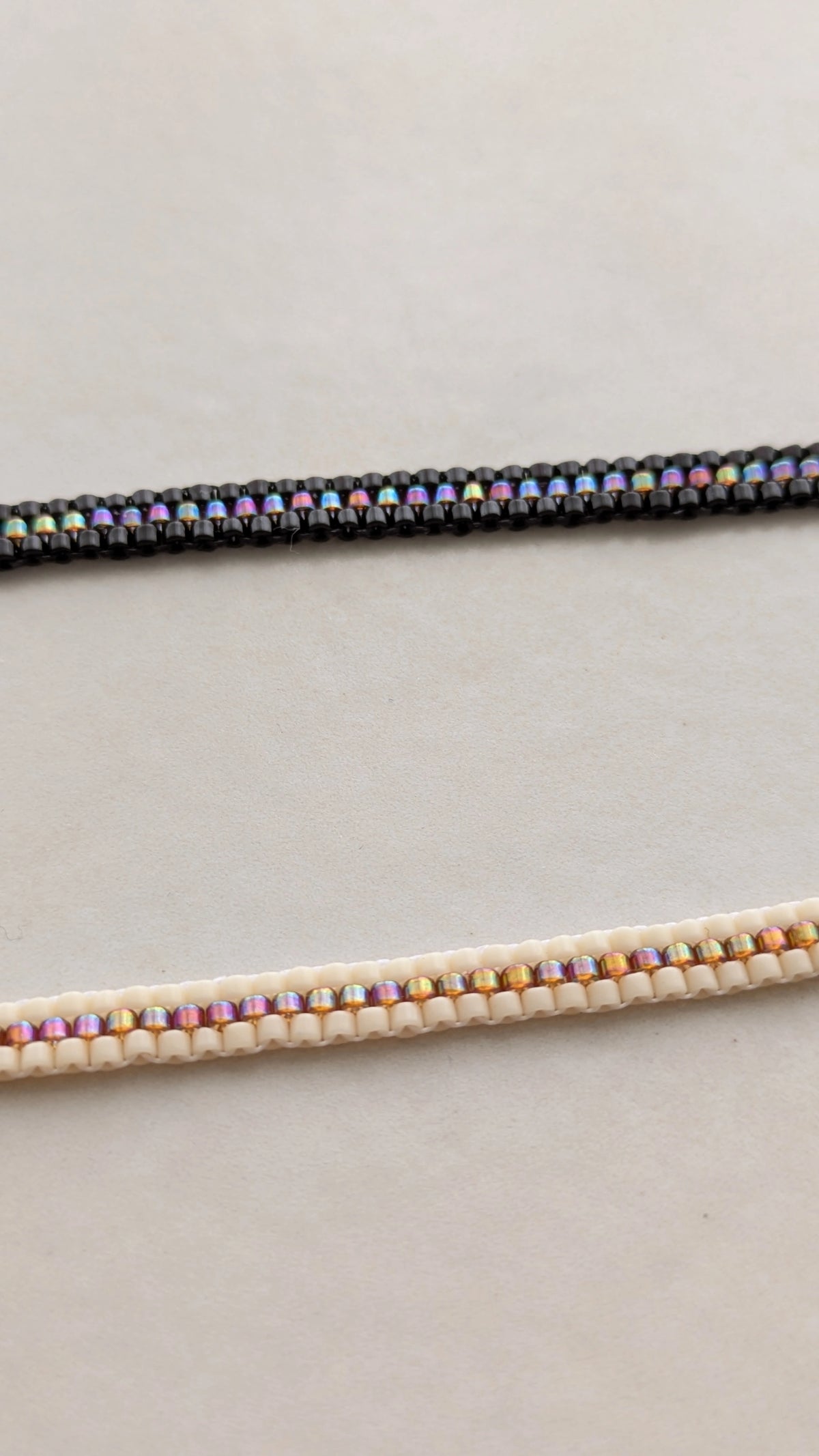 Vue détaillée sur deux bracelets . Un est noir l'autre crème, tous deux présentent une rangée de perles couleur arc en ciel en leur centre.