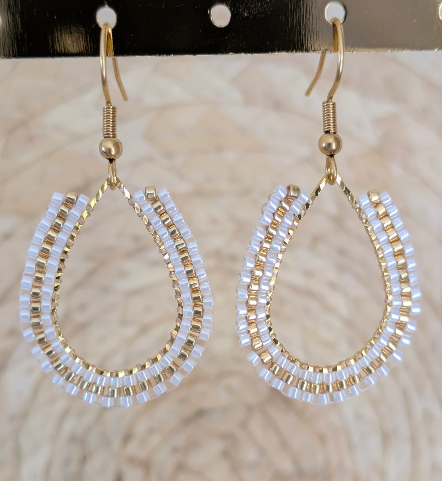 Boucles d'oreilles artisanales tissées sur goutte en métal doré. Gros plan sur les perles blanches et dorées.