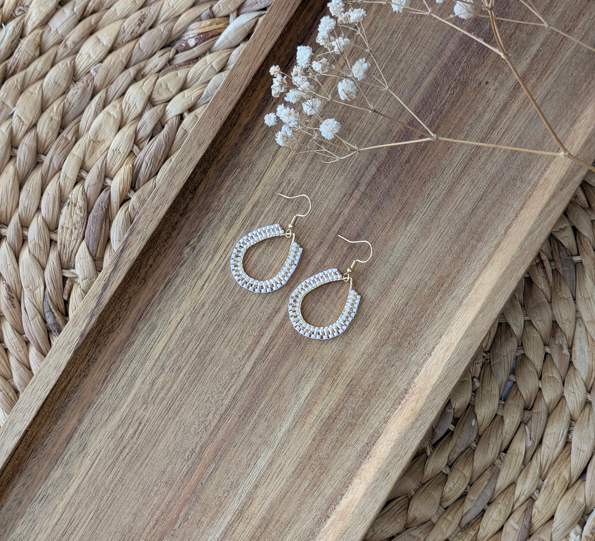 Boucles d'oreilles perlées blanches et dorées style bohème posées sur plat en bois.