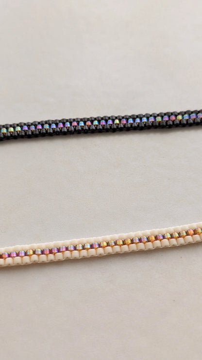 Vue détaillée sur deux bracelets . Un est noir l'autre crème, tous deux présentent une rangée de perles couleur arc en ciel en leur centre.